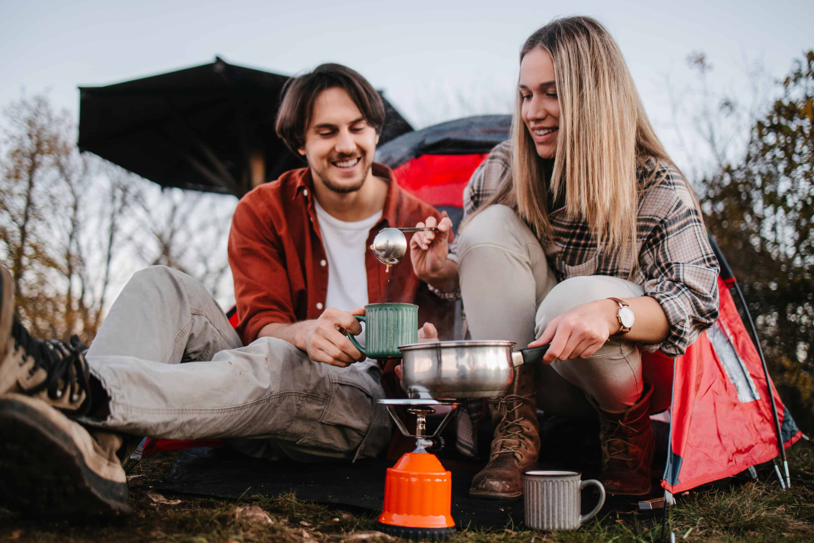Junges Paar sitzt vor einem Zelt und serviert Essen vom Campingkocher.