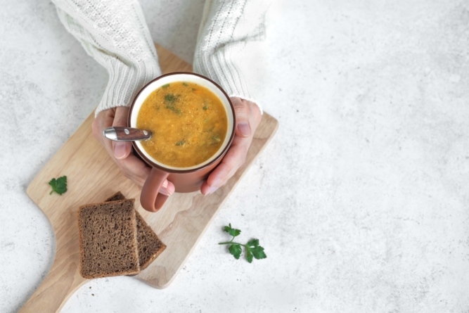 Hände wärmen sich an einer Tasse gefüllt mit heißer Suppe und Brot als Beilage.