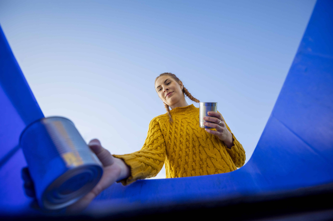 Junge Frau in gelbem Pullover entsorgt Konservendosen in einer blauen Tonne.