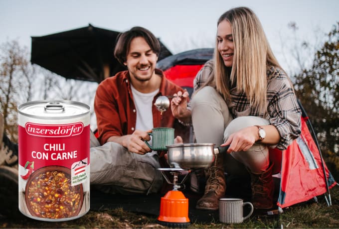 Junges Paar sitzt vor einem Zelt und serviert Essen vom Campingkocher und Chili con Carne.