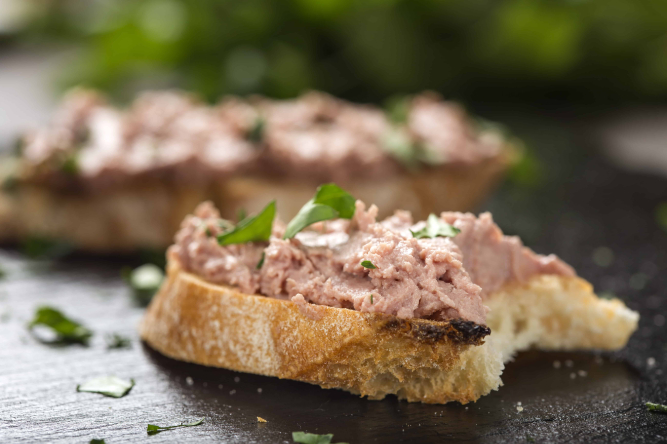 Knusprige Baguettescheibe mit Leberaufstrich auf schwarzem Holzbrett, garniert mit Petersilie.