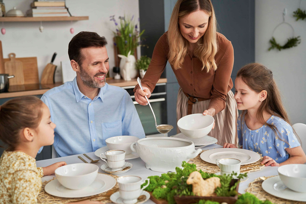 Vater und Kinder sitzen fröhlich an gedecktem Tisch, während die Mutter ihnen Suppe mit einer Kelle serviert.