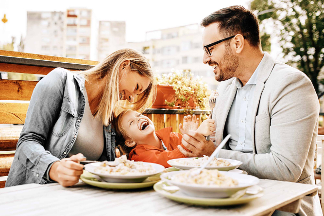 Mutter, Vater und Sohn sitzen lachend auf einer Terrasse und essen eine warme Mahlzeit.